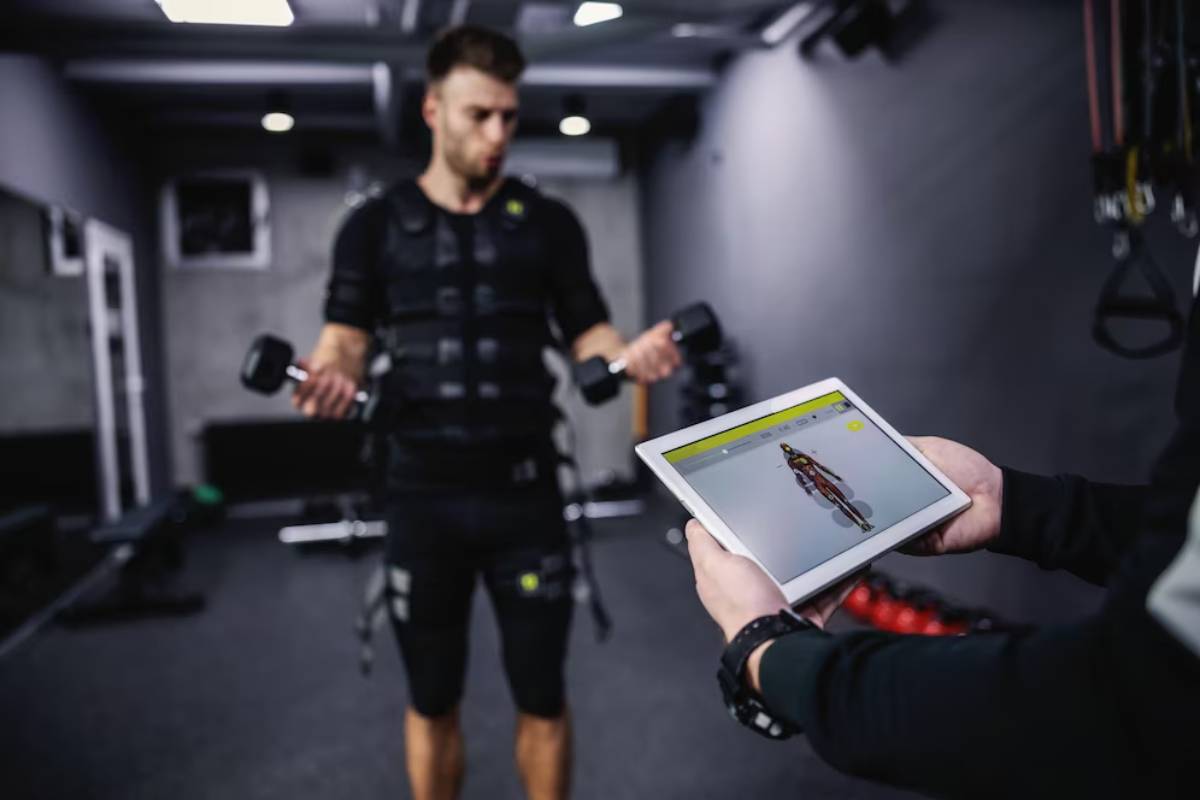 Man in fitness gear using electrode muscle stimulation while looking at a digital tablet displaying body metrics in a gym.