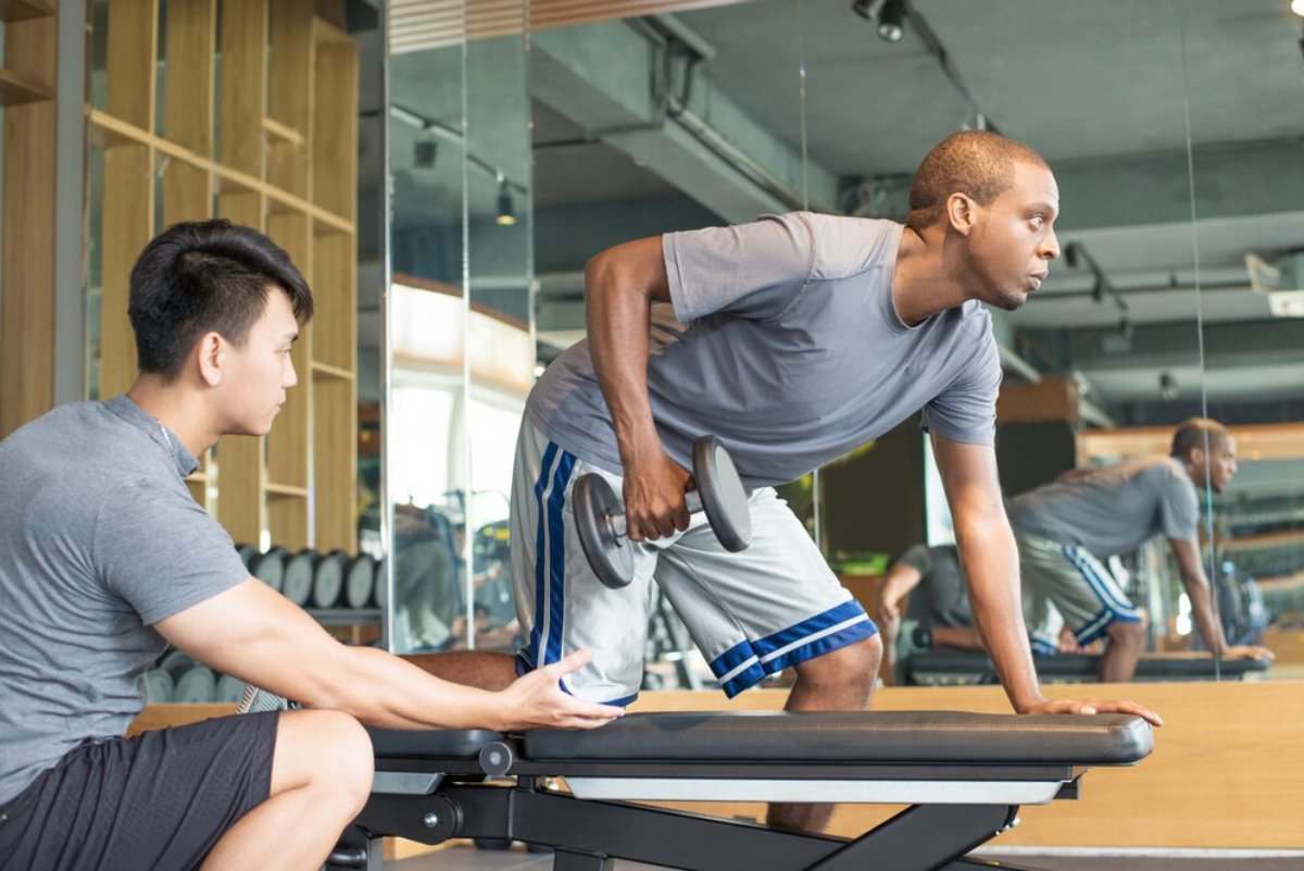A personal trainer assists a man lifting dumbbells in a gym with reflective mirrors.