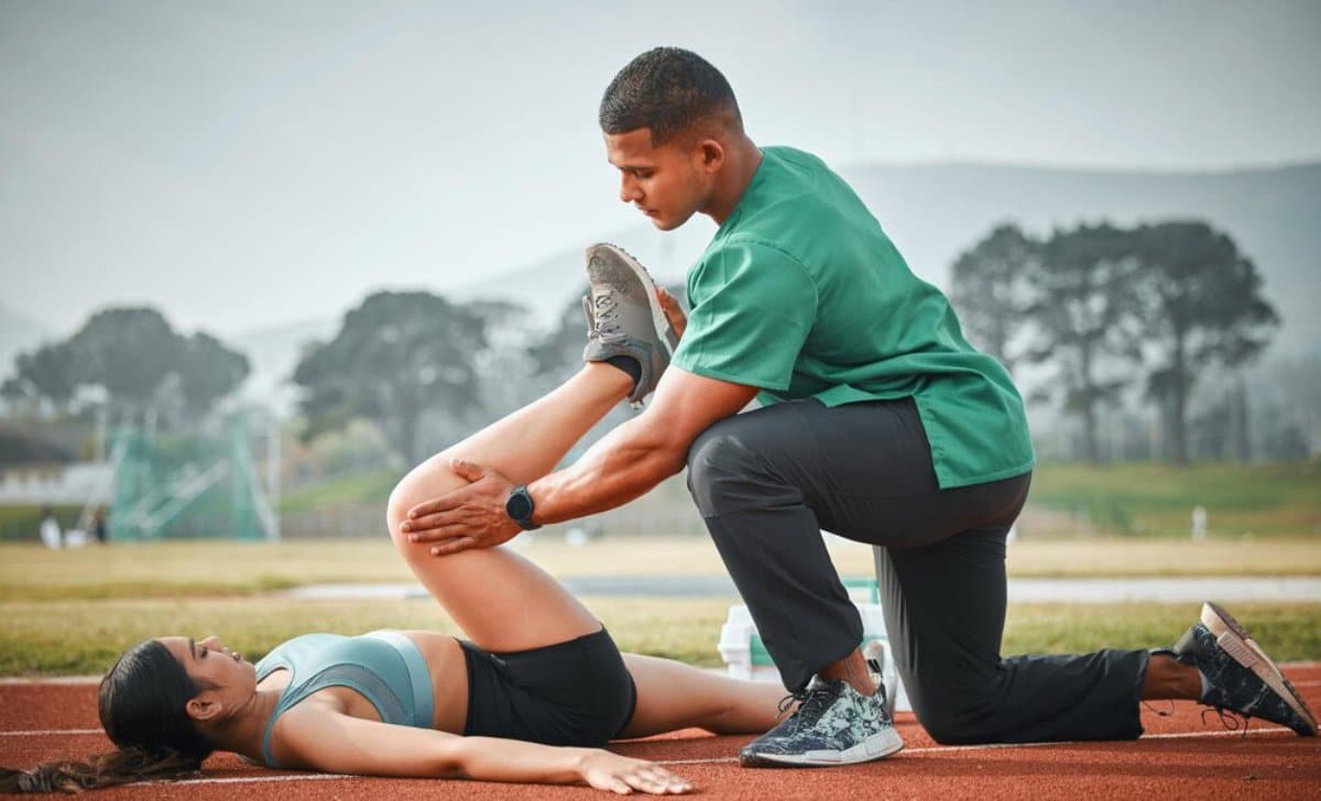 A male trainer stretching a female athlete's leg on a running track outdoors.