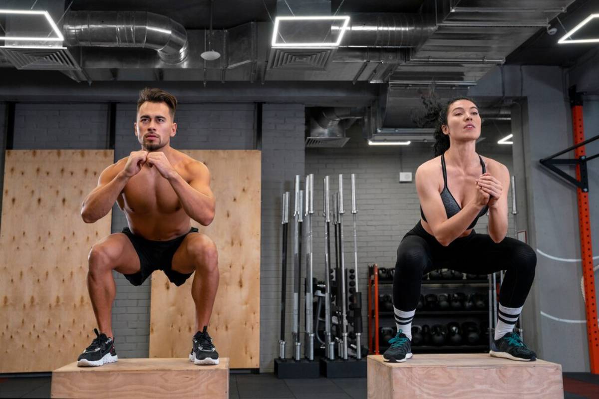 A man and a woman performing squats on wooden boxes in a gym.