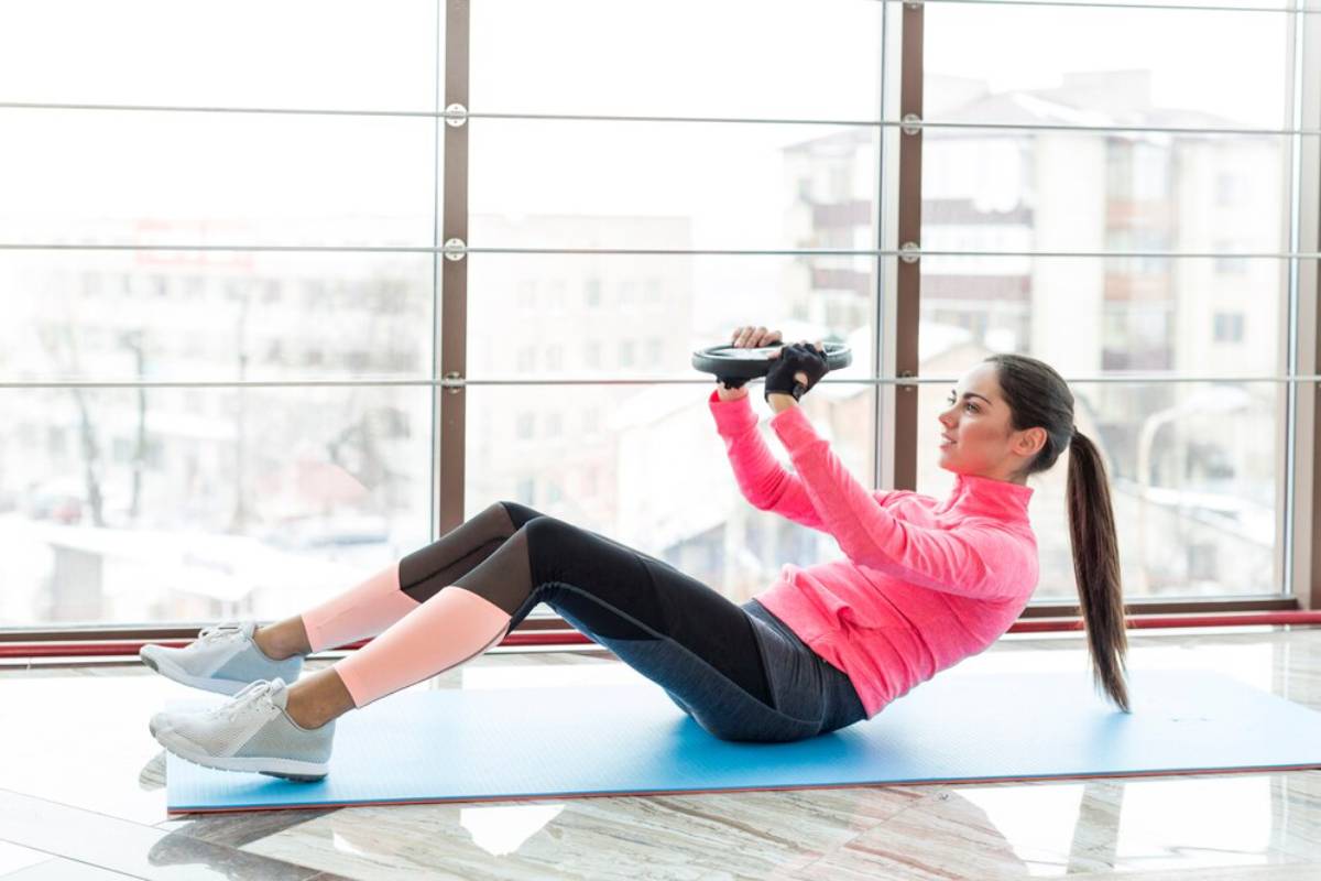 Woman in pink shirt exercising with a weighted plate in a brightly lit gym with large windows.