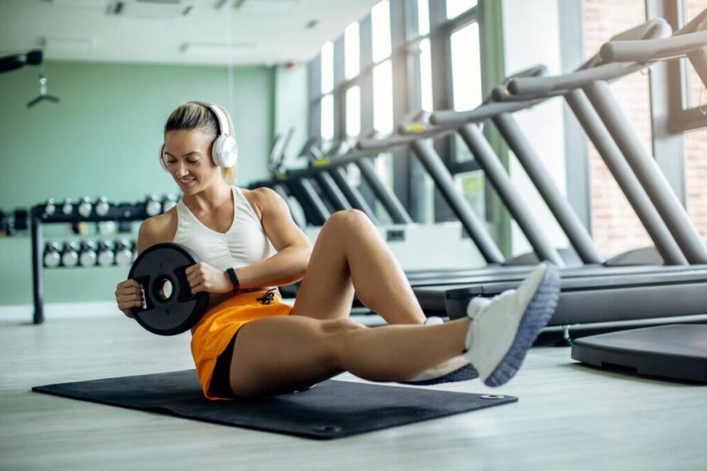 Young woman in gym clothes using a weight plate while sitting on a mat in a fitness center.
