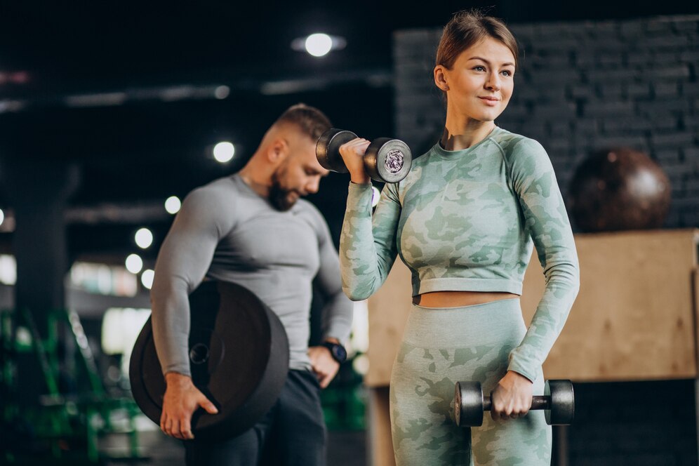 Two people exercising in a gym, woman with dumbbells and man lifting a weight plate, both wearing camo workout gear.