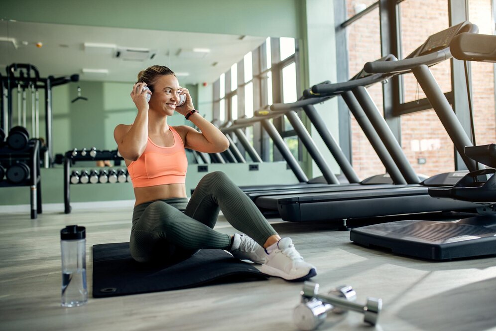 A young woman sitting on a gym floor, putting on headphones, surrounded by exercise equipment.