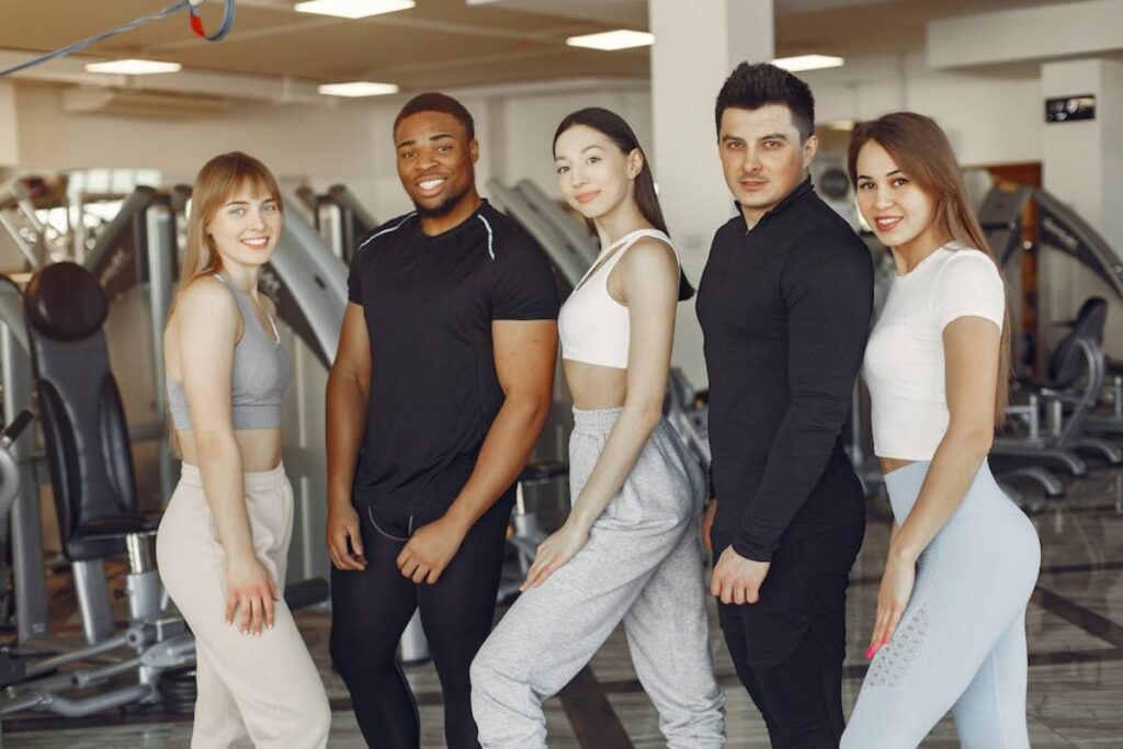 Five diverse young adults posing together in a gym, smiling at the camera.