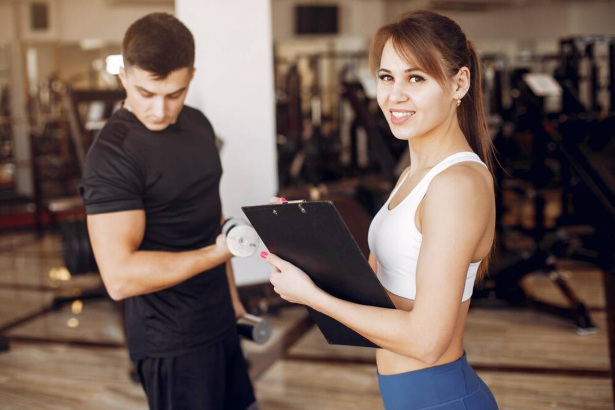 Young woman in fitness attire holding a clipboard and smiling at the camera while a young man lifts weights in a gym.