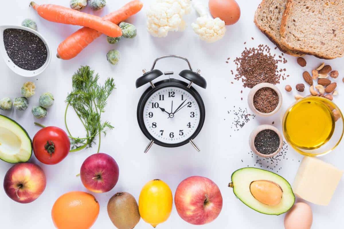 Alarm clock surrounded by various healthy foods like fruits, vegetables, nuts, and grains on a white background.