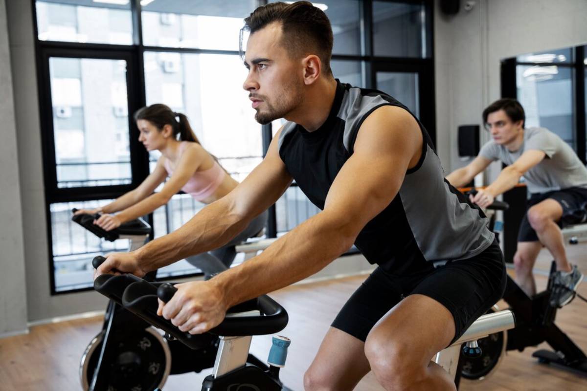 Three focused people using stationary bikes in a fitness class at a gym with large windows.