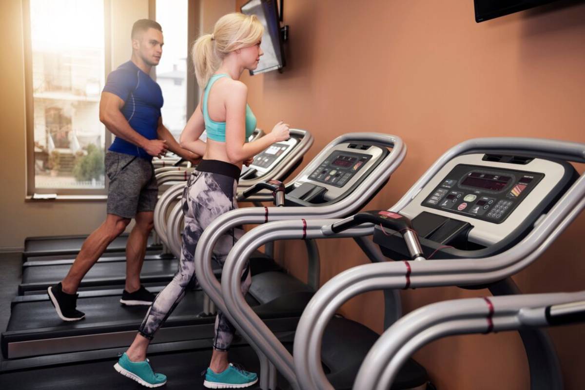 A man and a woman using treadmills in a modern gym with sunlight filtering in.