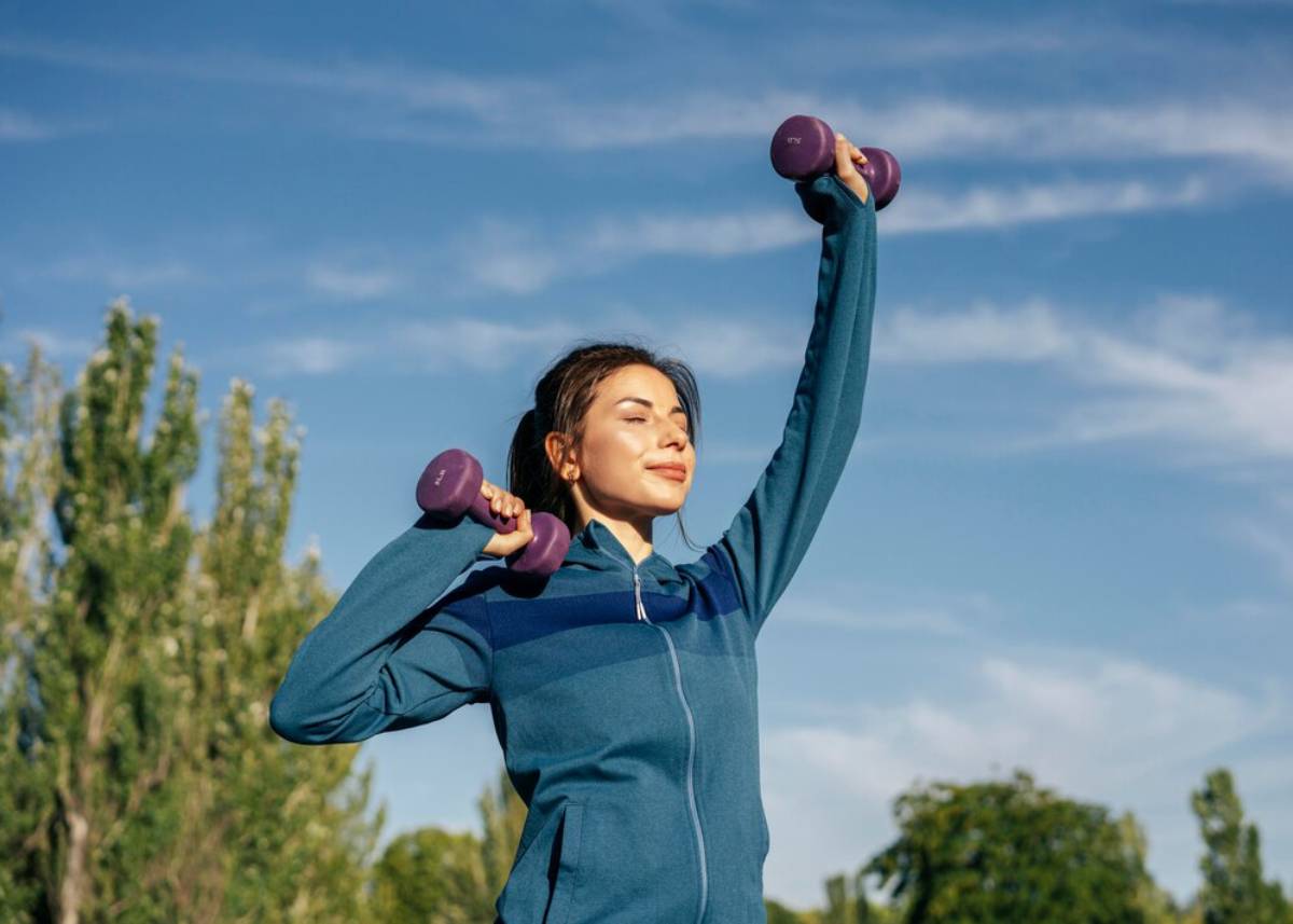 A woman exercises outdoors lifting purple dumbbells under a clear blue sky.