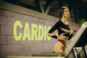 A young woman jogs on a treadmill in a gym with a neon "CARDIO" sign on the wall.