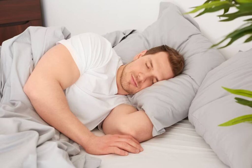 Man sleeping peacefully in a well-lit bedroom, with white bedding and a plant in the background.