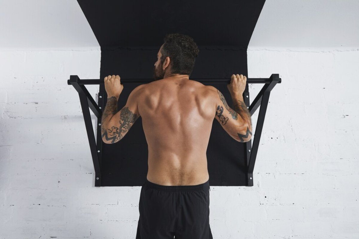 A muscular man with tattoos doing pull-ups on a bar in a white room.
