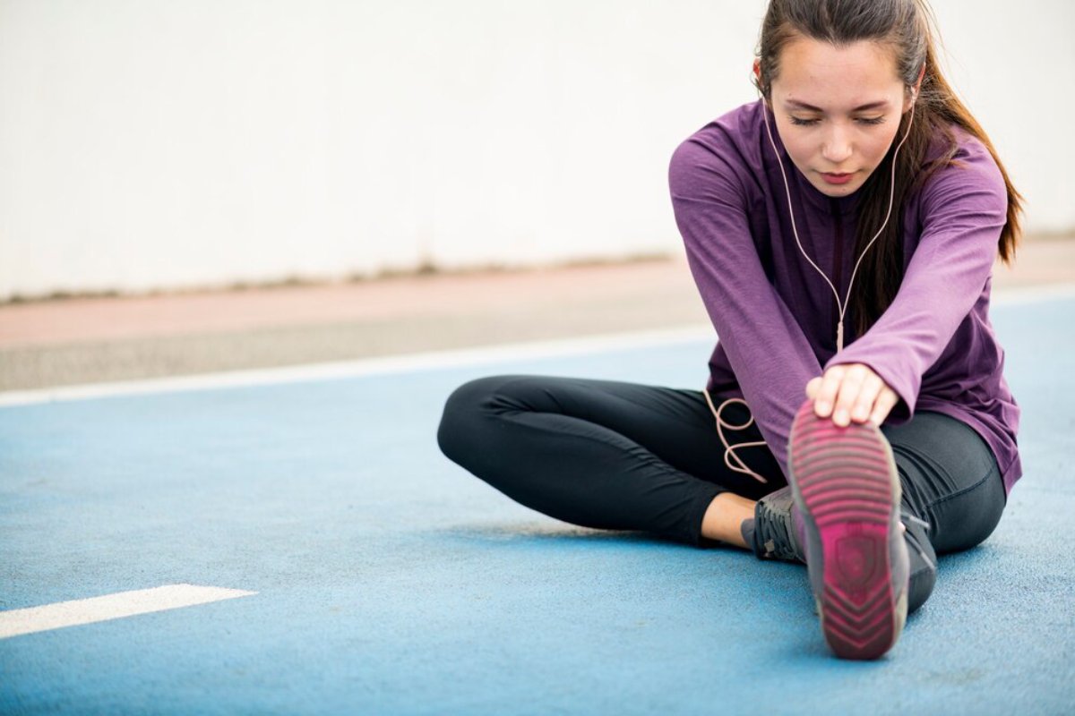 Woman in purple top stretching on a blue track, reaching towards her foot while wearing headphones.