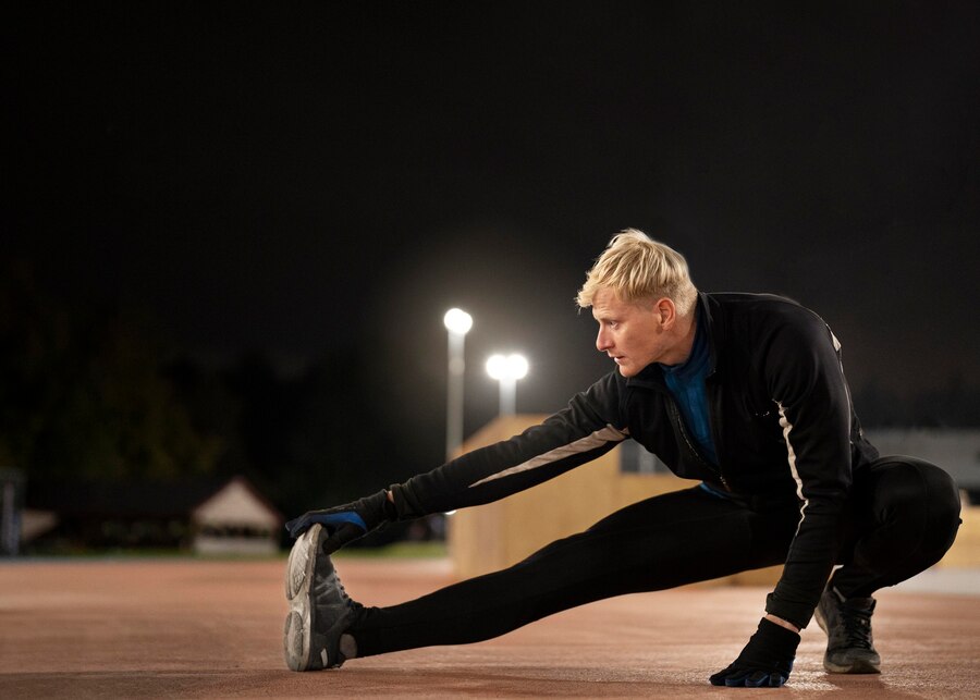 Man stretching on a track at night under bright lights.